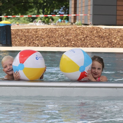 Drei Kinder plantschen mit Wasserbällen in einem Schwimmbecken.