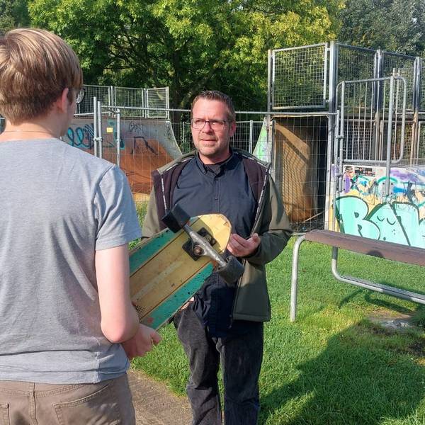 Ein Jugendlicher mit Skateboard in den Händen unterhält sich mit einem Mann vor einer Skateanlage.