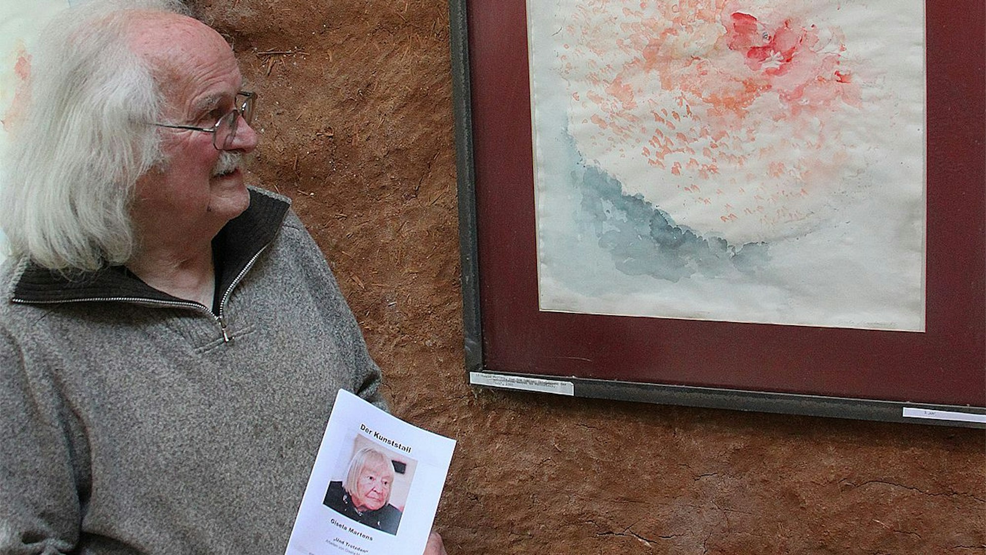 Wolfgang Martens steht an einem Gemälde, das an einer rot-braunen Wand hängt. Der Mann hat weißes Haar und trägt einen grauen Pullover.