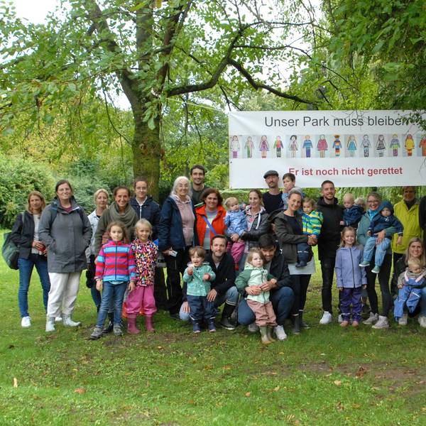 Männer, Frauen und Kinder stehen vor einem Banner, das in einem Park zwischen zwei Bäumen hängt.