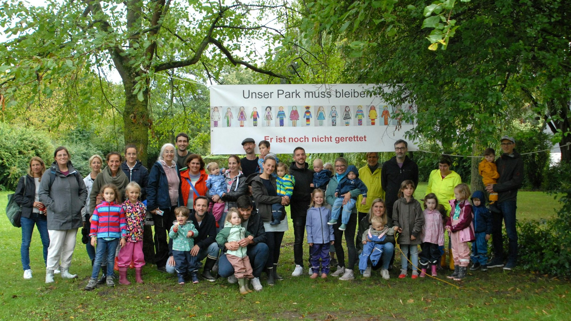 Männer, Frauen und Kinder stehen vor einem Banner, das in einem Park zwischen zwei Bäumen hängt.
