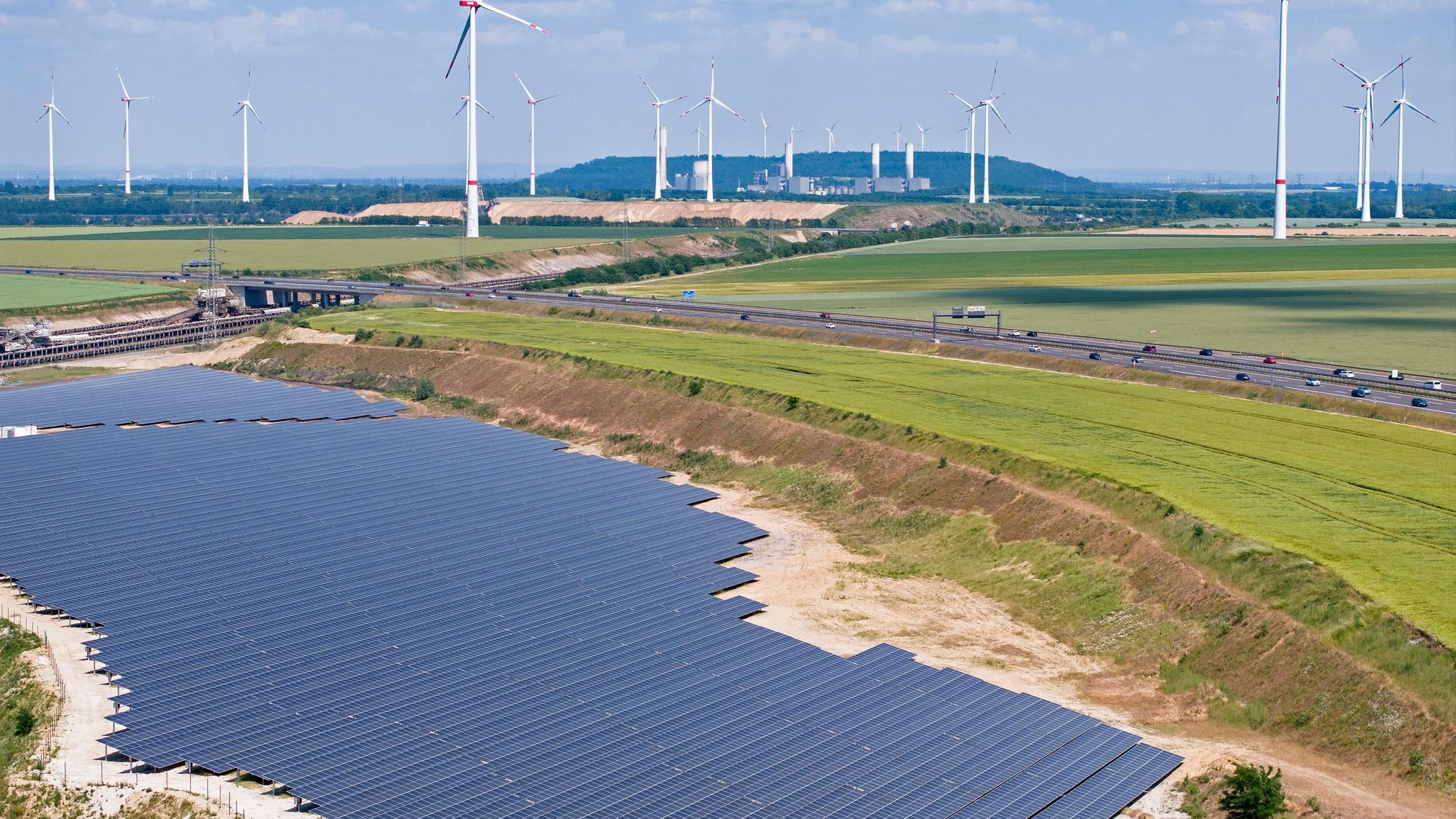 Aus der Luft ist eine große Photovoltaikanlage zu sehen, im Hintergrund sind eine Autobahn, Äcker und Windräder.