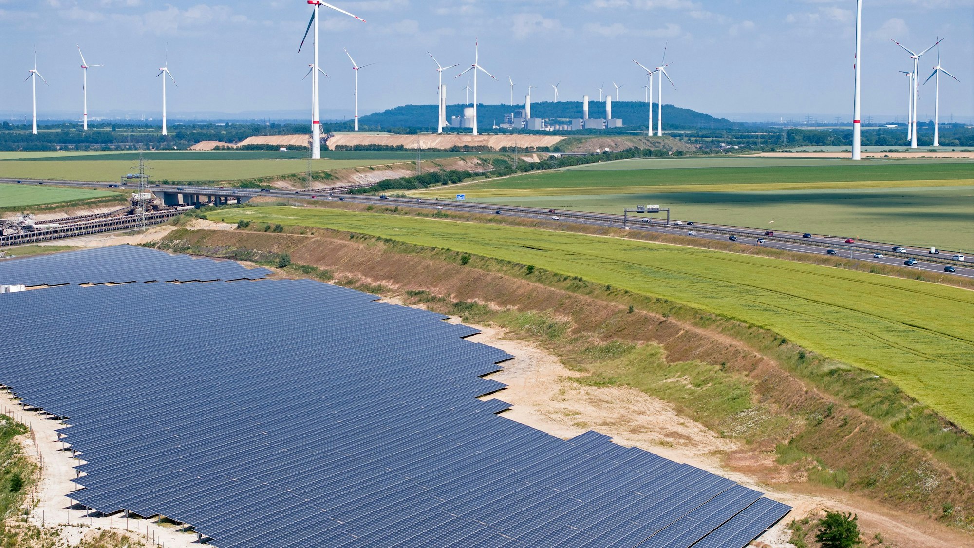 Aus der Luft ist eine große Photovoltaikanlage zu sehen, im Hintergrund sind eine Autobahn, Äcker und Windräder.