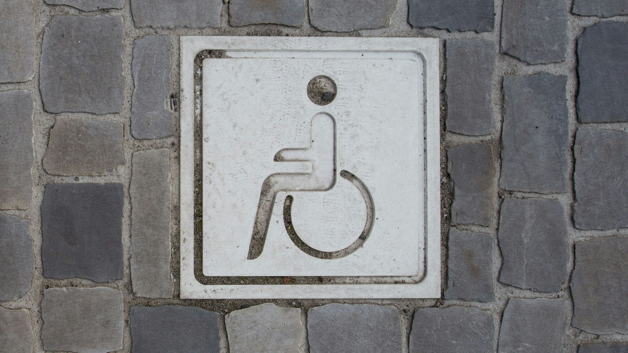 Auf einem Boden aus Pflastersteinen ist ein Piktogramm von einer Person in einem Rollstuhl.