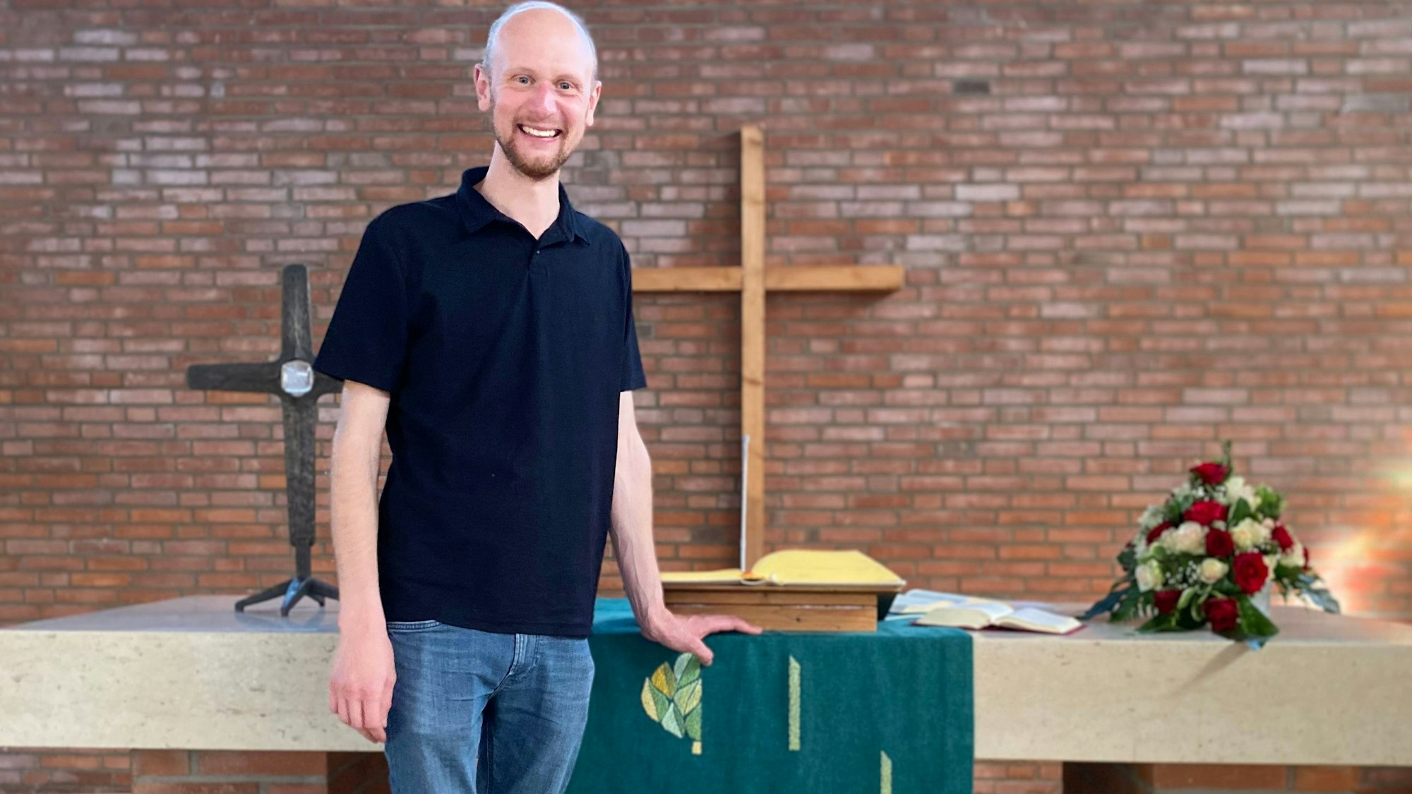 Diakon Jan Simons steht vor dem Altar der Evangelischen Kirche in Weilerswist. An der Wand hängt ein hölzernes Kreuz. Auf dem Altar liegen Blumen und die Bibel.