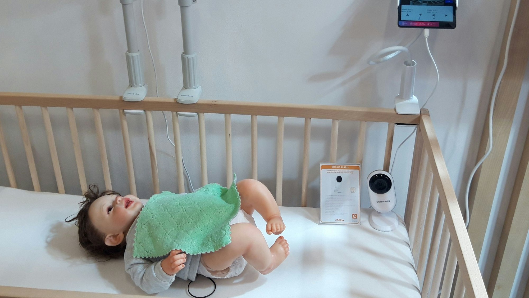 In einem Gitterbett liegt eine Babypuppe, über ihr hängt eine Kamera, daneben zeigt ein Tablett ein Bild von der Situation im Bett an.
