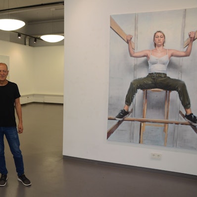 Ein Mann im schwarzen T-Shirt, Jeans und dunklen Sneakern steht im Ausstellungsraum. Links und rechts hängen Bilder, die er gemalt hat.