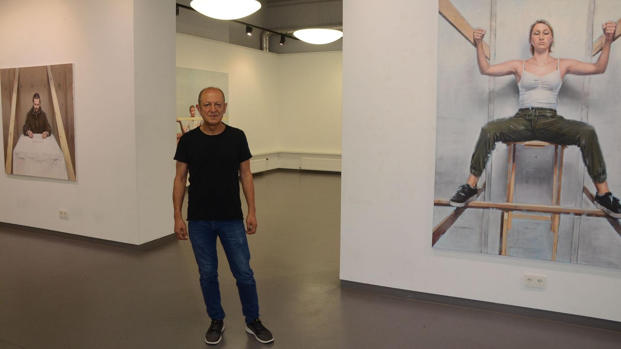 Ein Mann im schwarzen T-Shirt, Jeans und dunklen Sneakern steht im Ausstellungsraum. Links und rechts hängen Bilder, die er gemalt hat.