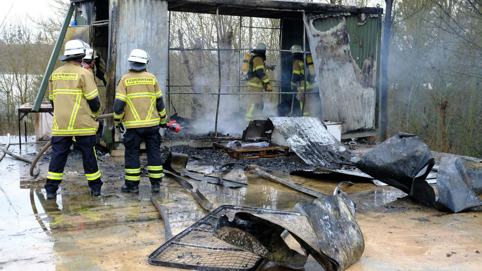 Feuerwehrleute stehen in Mechernich vor und in einem Wohncontainer, der durch einen Brand völlig zerstört worden ist.