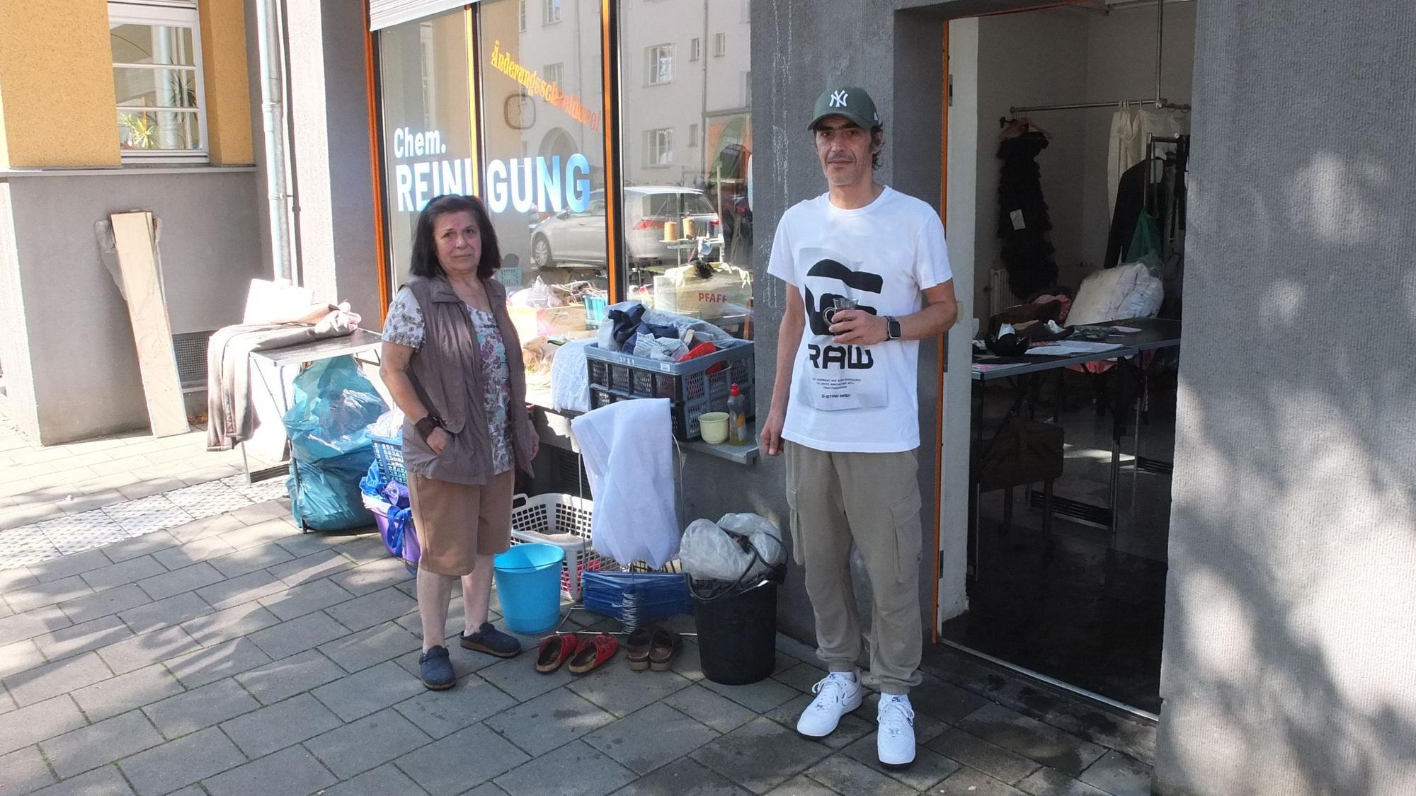 Ein Mann und eine Frau stehen vor einem Laden, die Tür steht offen, Säcke und Putzeimer stehen davor.