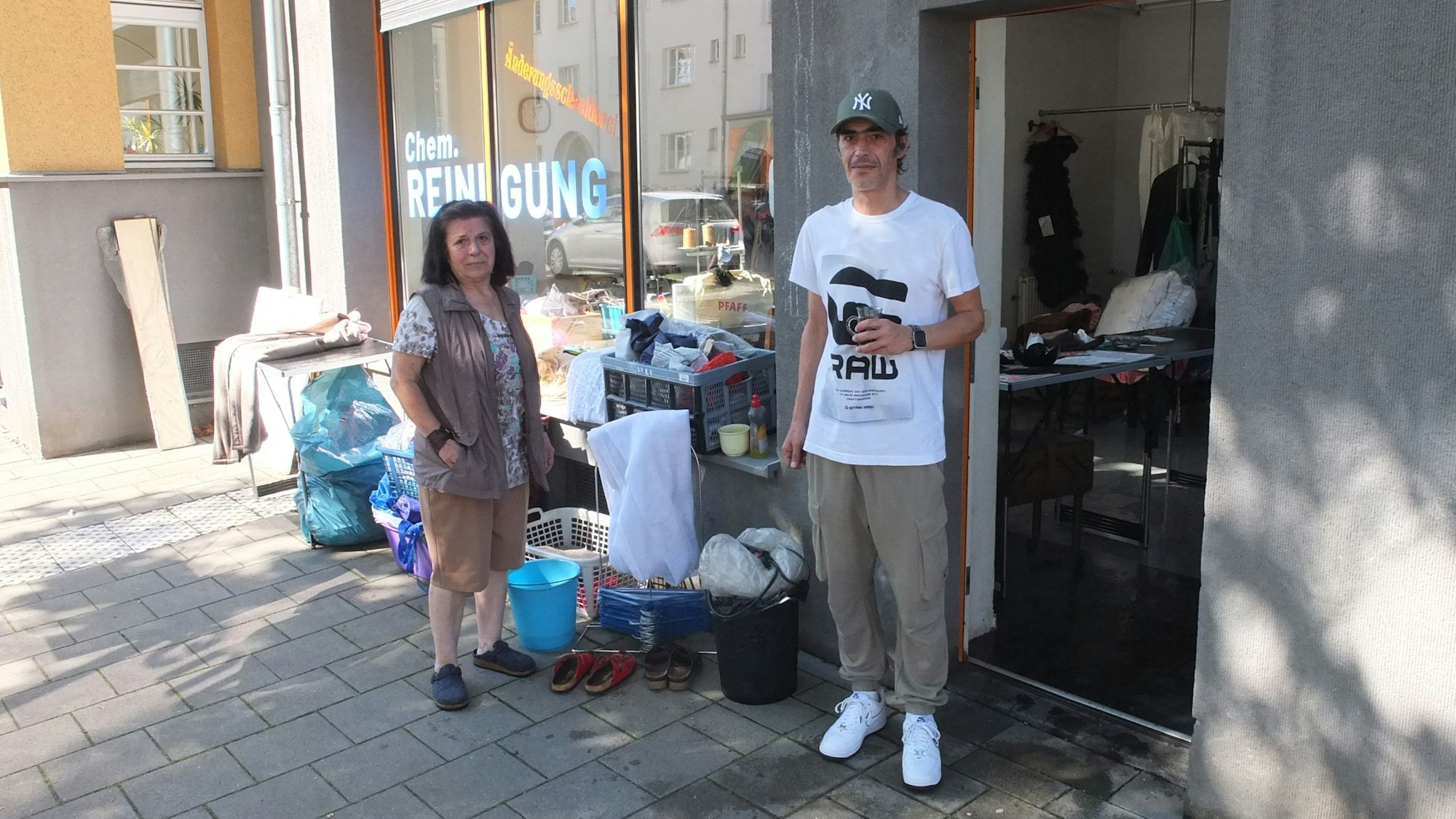 Ein Mann und eine Frau stehen vor einem Laden, die Tür steht offen, Säcke und Putzeimer stehen davor.