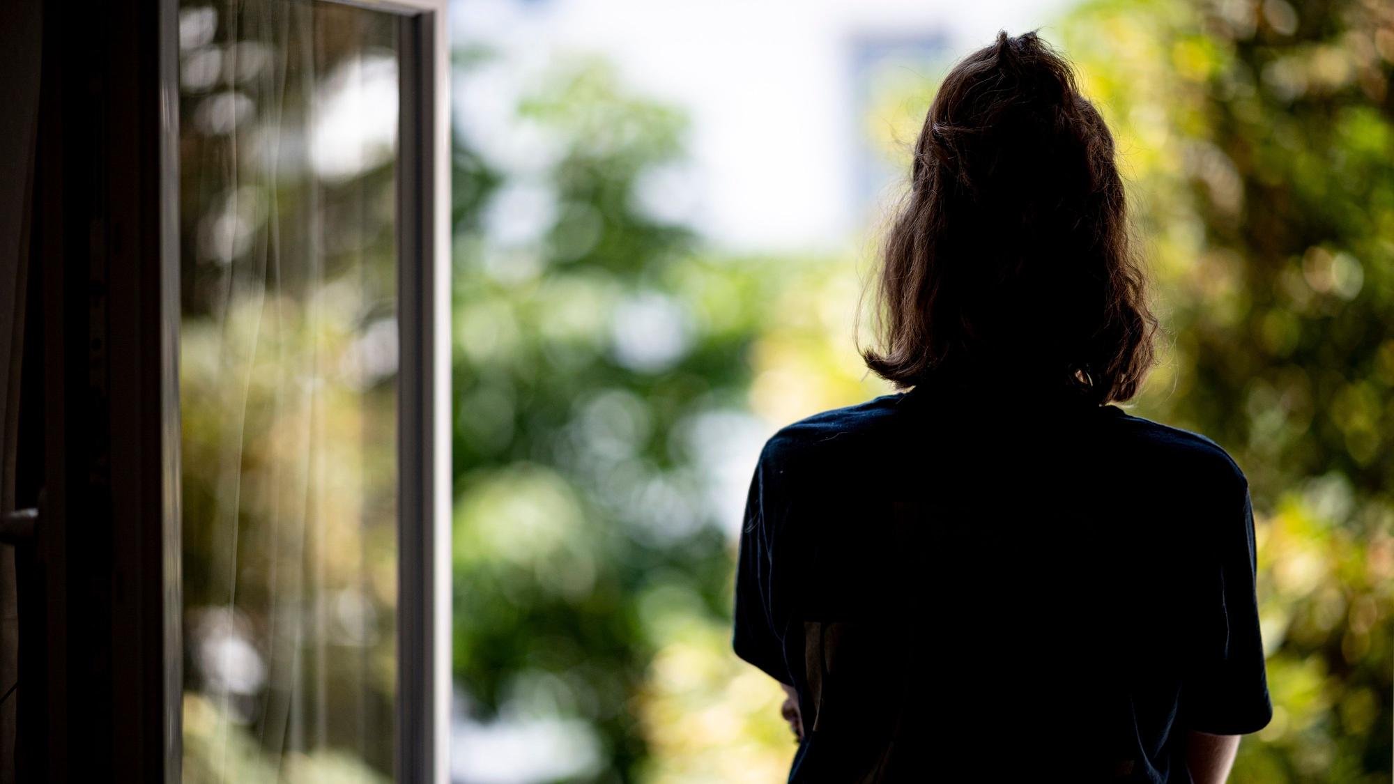 Eine junge Frau steht in ihrer Wohnung an einem Fenster.