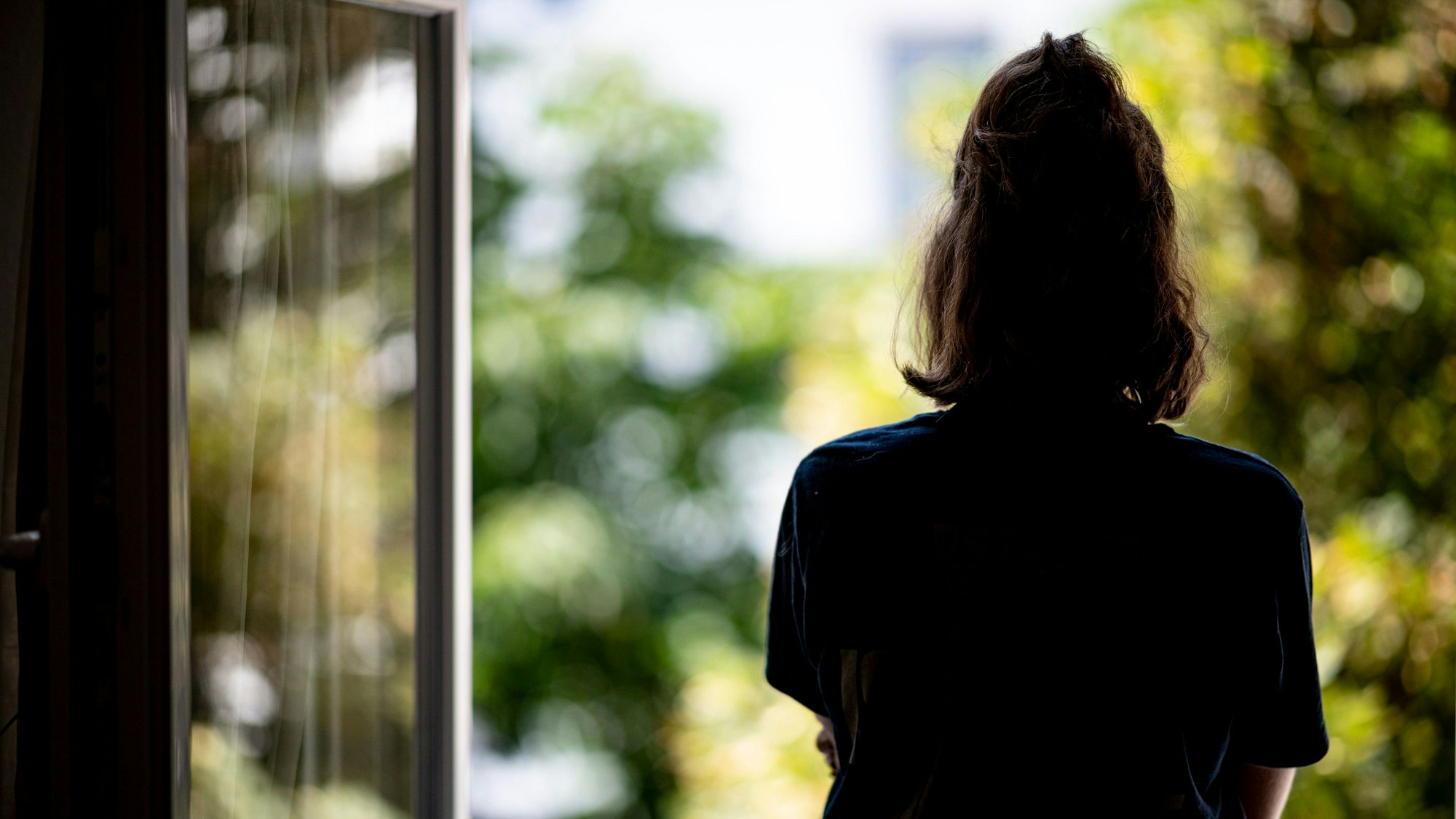 Eine junge Frau steht in ihrer Wohnung an einem Fenster.