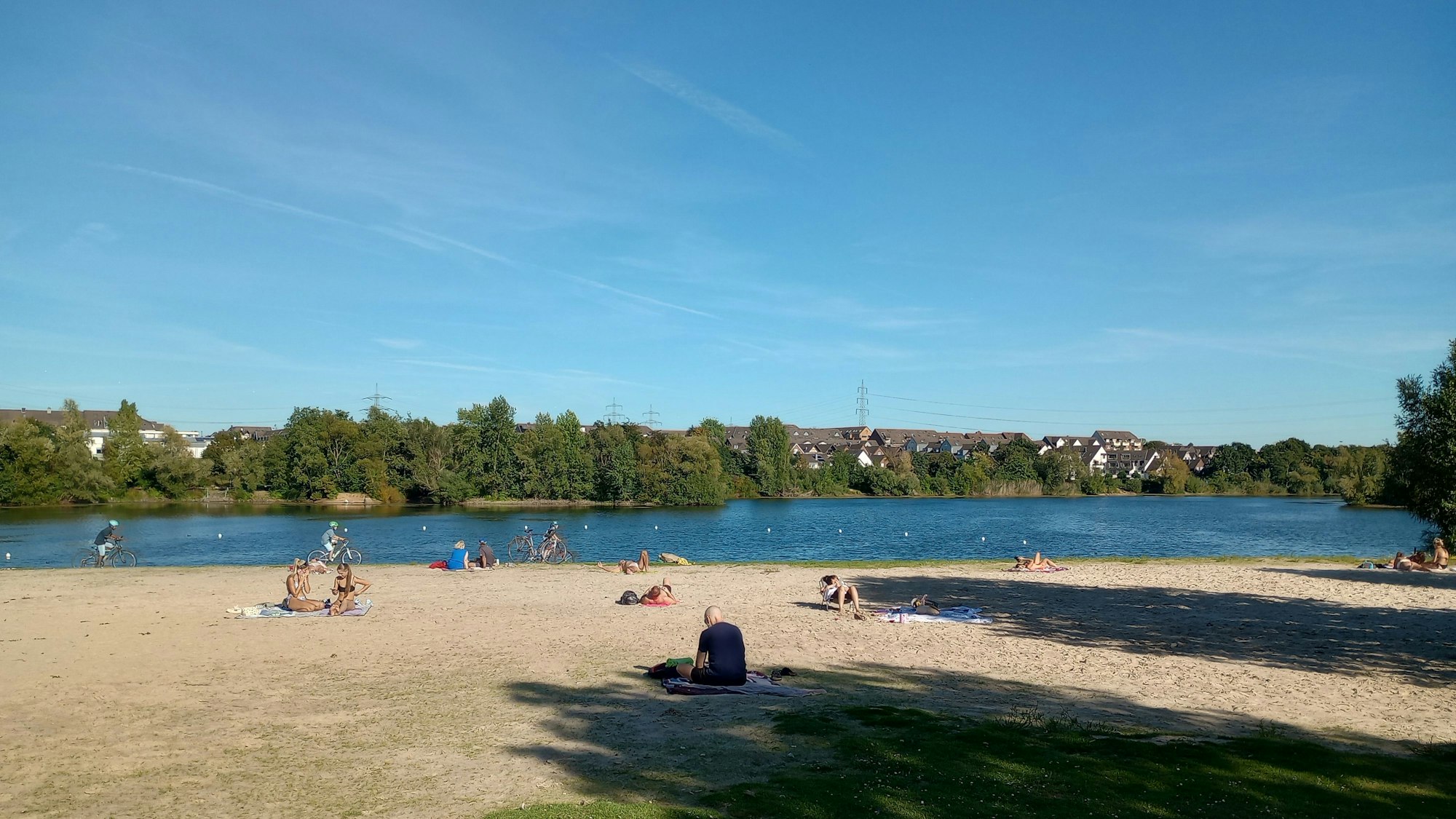 Menschen sitzen am sandigen Ufer eines Baggersees.