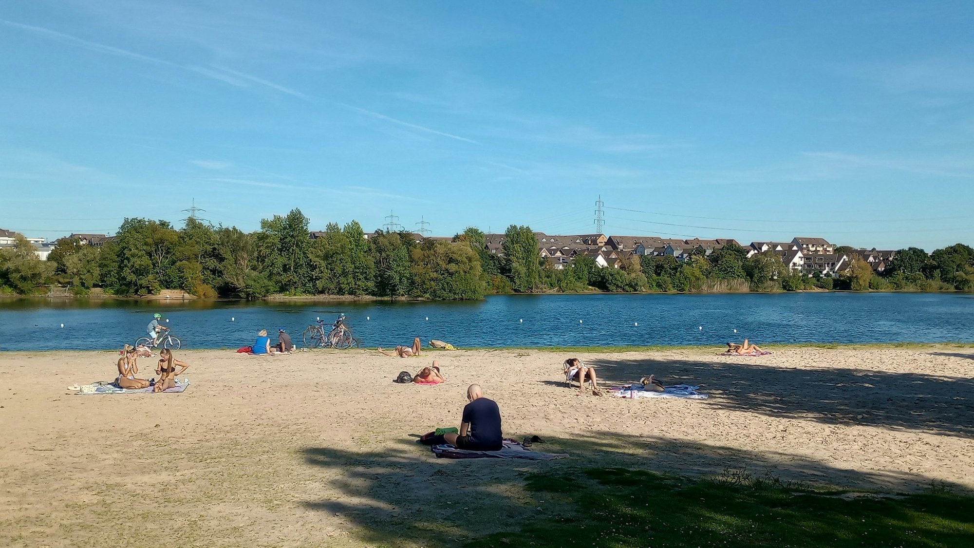 Menschen am sandigen Ufer eines Badesees. Im Hintergrund sind Bäume und Büsche am anderen Ufer zu sehen, außerdem die Dächer von Häusern.