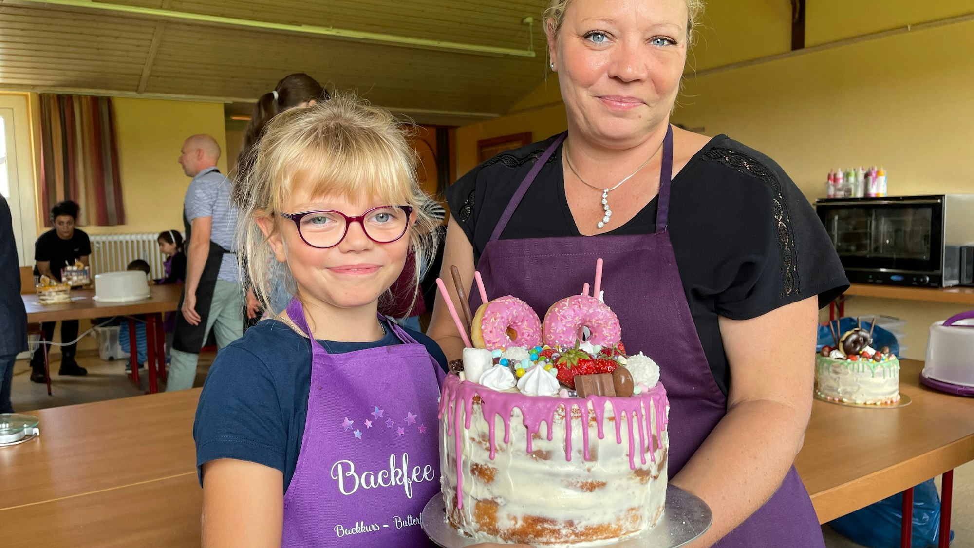 Ein blondes Mädchen und eine blonde Frau, beide in lila Schürzen, halten einen Naked Cake mit pinkfarbenen Donuts und rosa Glasur.
