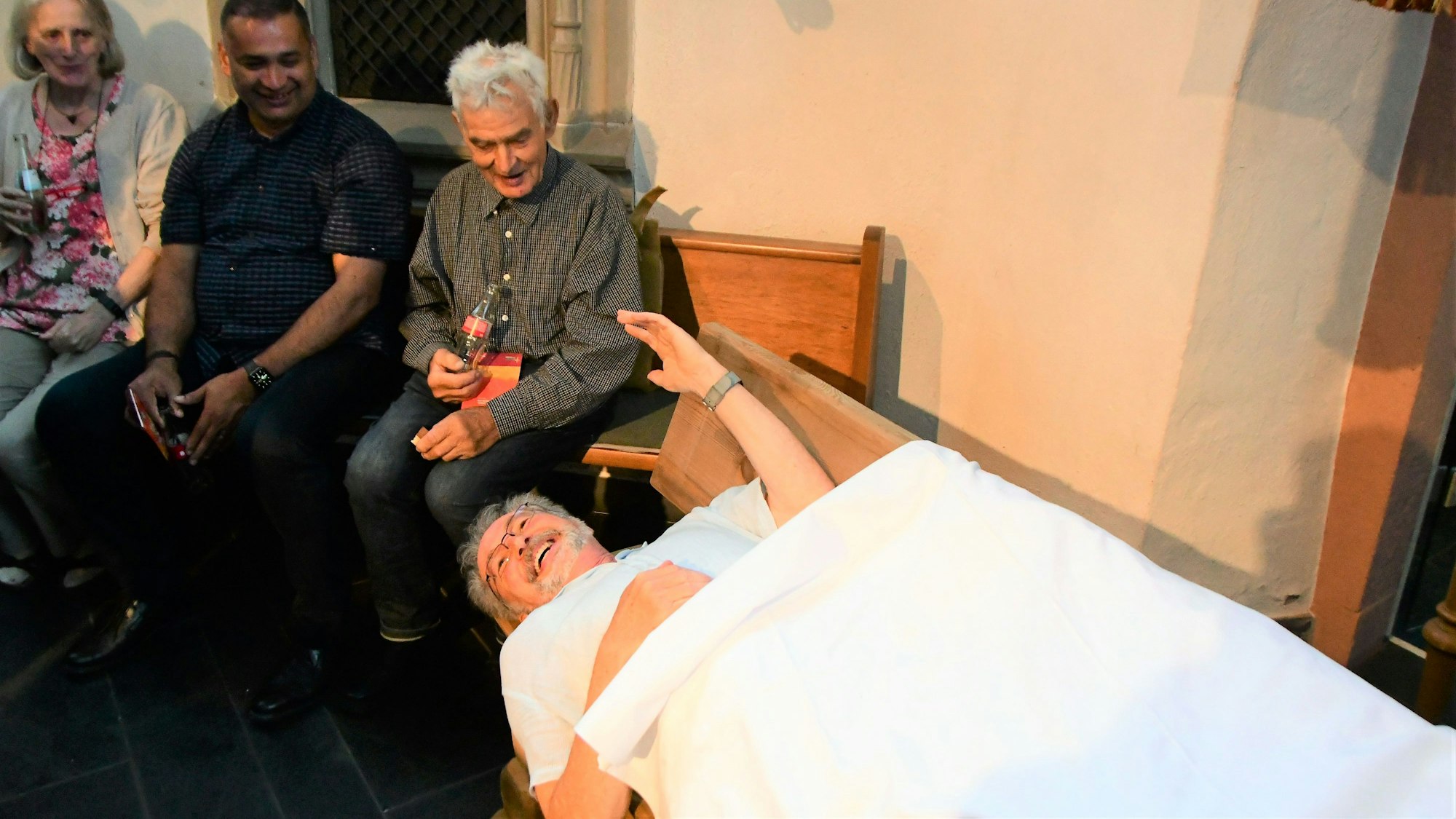 Pfarrer Ludwig Pützkaul liegt lachend und zugedeckt auf der ihm geschenkten Bank. Weitere Gäste sind amüsiert.