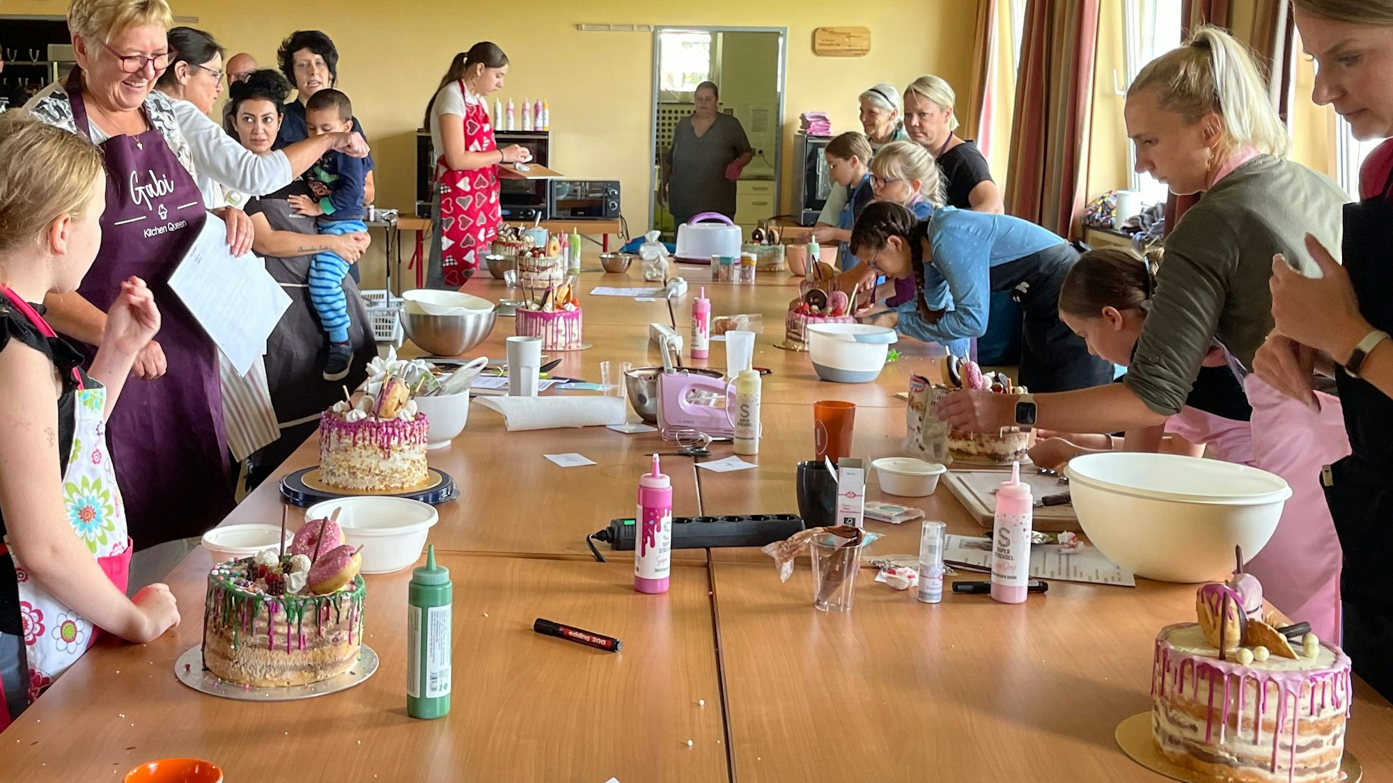 Viele lachende Frauen und Kinder stehen an langen Tischen, auf denen mehrstöckige, reich verzierte Torten, Schüsseln, Fläschchen und Mixer stehen.