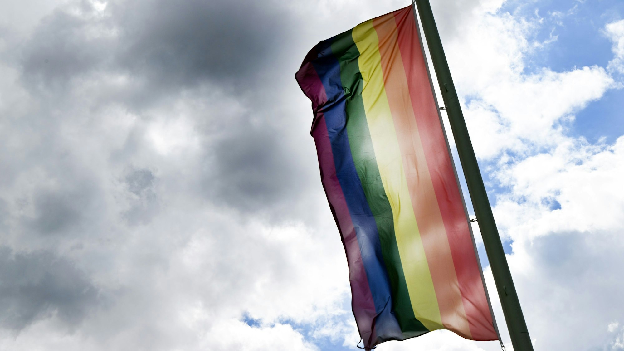 Eine Regenbogenflagge weht vor einem bewölkten Himmel. (Symbolbild)