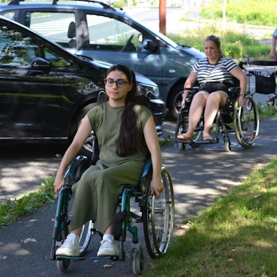 Junge Menschen in Rollstühlen passieren einen engen Gehweg.