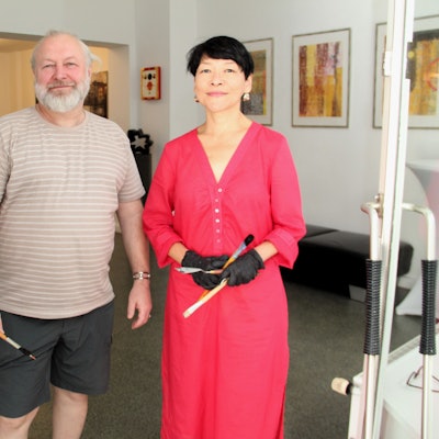 Ein Mann mit grauem Haar und Bart stehe neben einer Frau im roten Kleid mit kurzem schwarzem Haar. Beide haben Pinsel in der Hand.