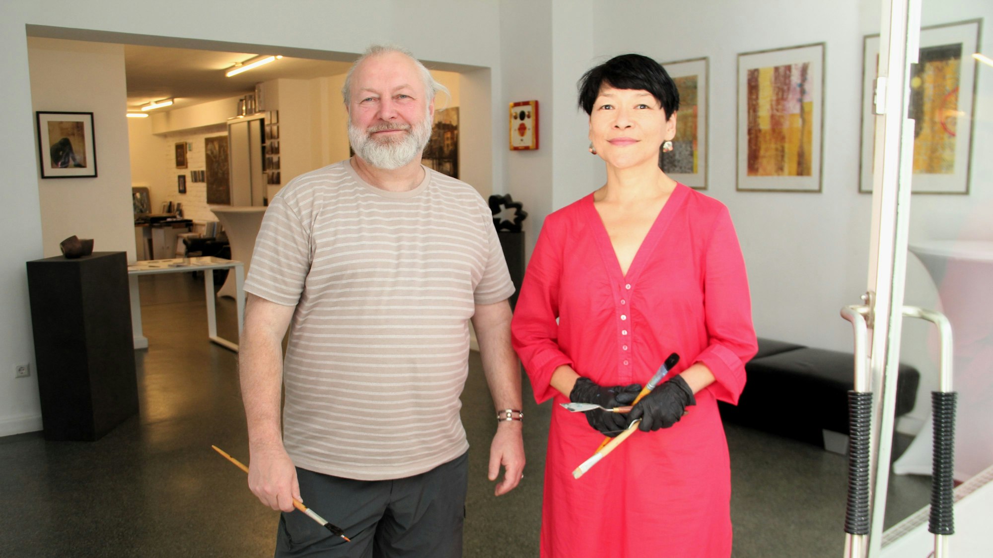 Ein Mann mit grauem Haar und Bart stehe neben einer Frau im roten Kleid mit kurzem schwarzem Haar. Beide haben Pinsel in der Hand.