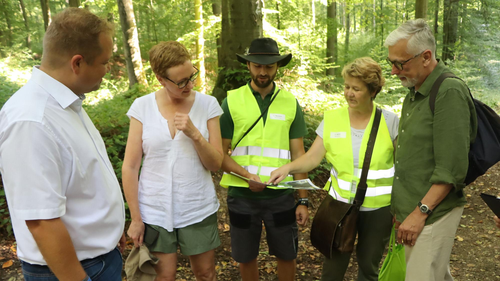 Eine Gruppe aus drei Männern und zwei Frauen, zwei Personen in gelben Warnwesten, schauen im Wald auf eine Karte.
