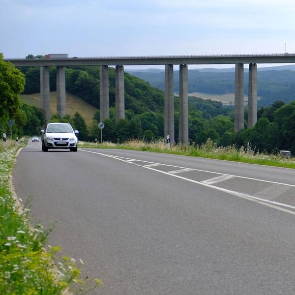 Das Bild zeigt die Talbrücke Zingsheimer Wald der Autobahn 1. Auf einer Landstraße, die unter der Brücke verläuft, sind zwei Autos unterwegs.