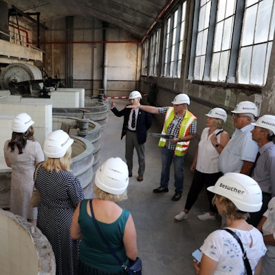 Menschen stehen in einer Halle und erhalten Erklärungen von einem Mann mit Warnweste.