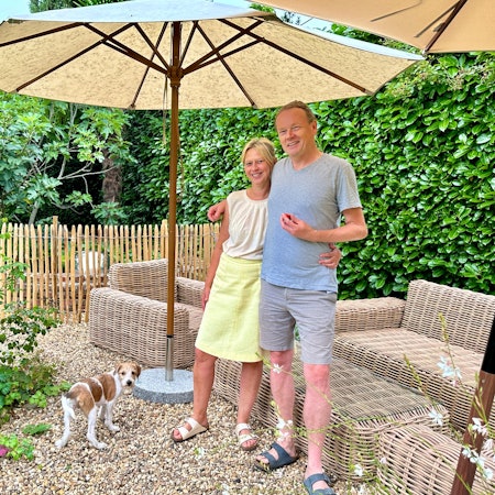 Ein Paar steht in einem Garten vor einer Draußen-Sitzgarnitur aus Korbgeflecht. Sie stehen unter einem Sonnenschirm, ein weißer Hund mit braunen Flecken schaut in die Kamera.
