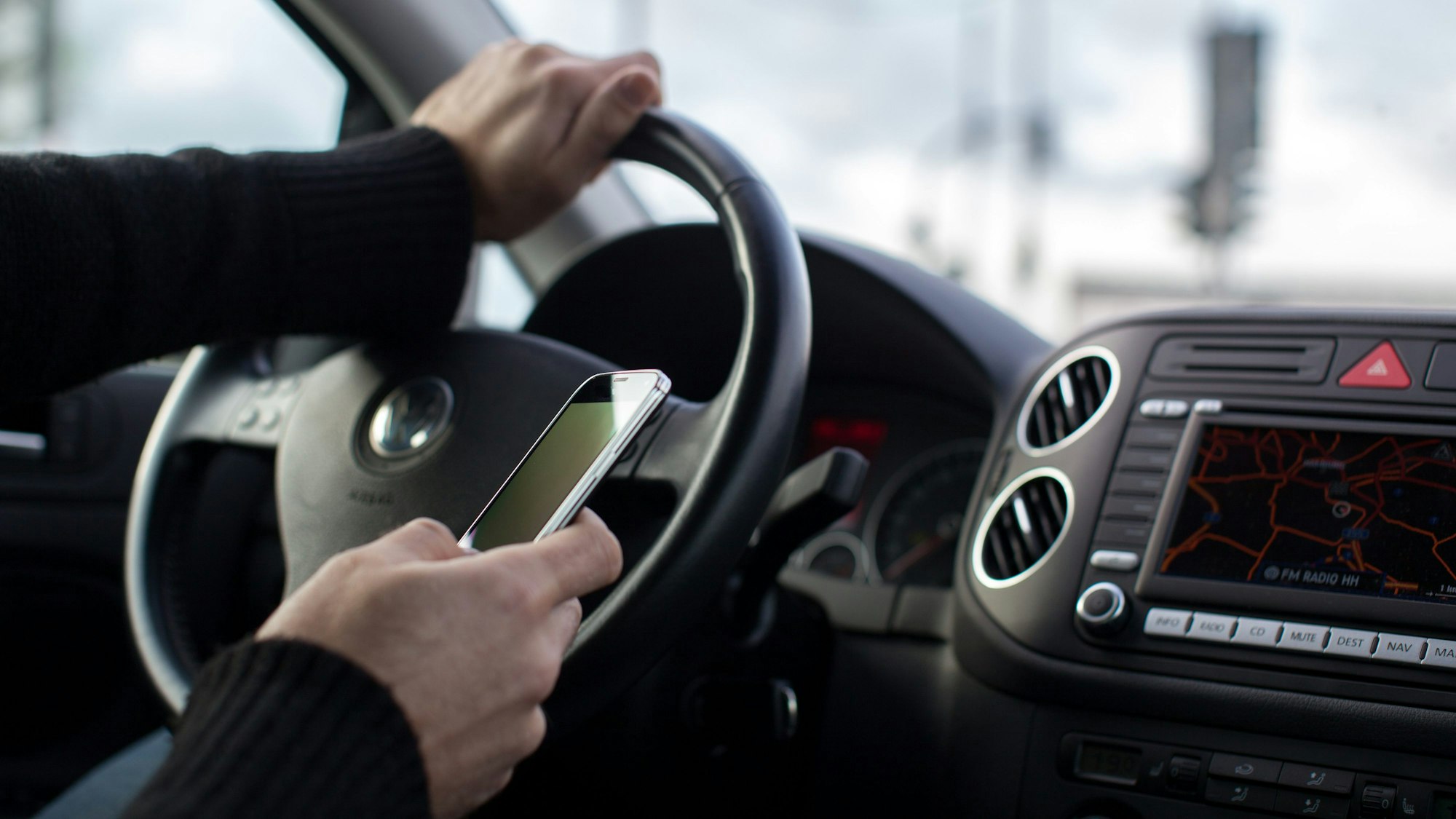 Ein Mann hat eine Hand am Steuer eines Autos und eine Hand am Smartphone.