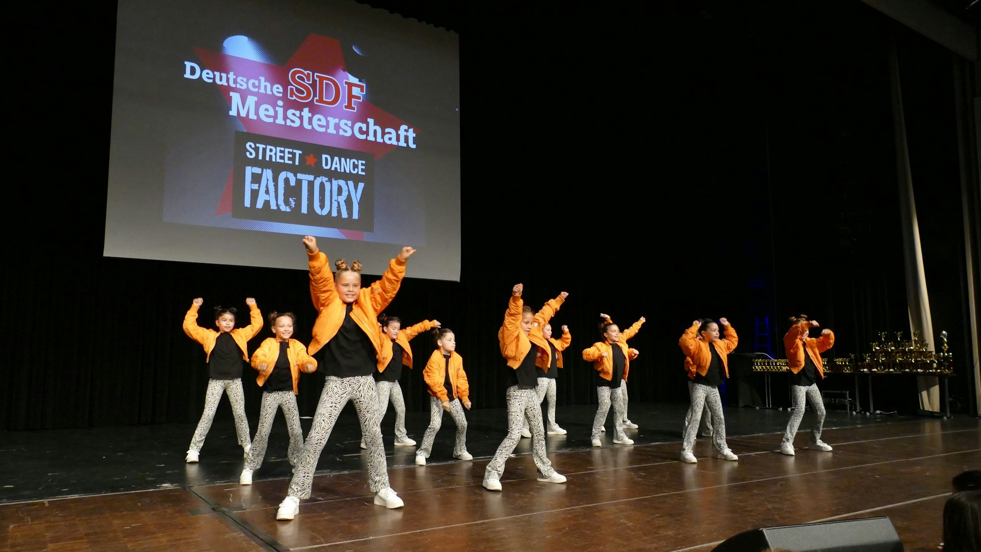 Eine Tanzgruppe in orangenen Jacken steht auf der Bühne.
