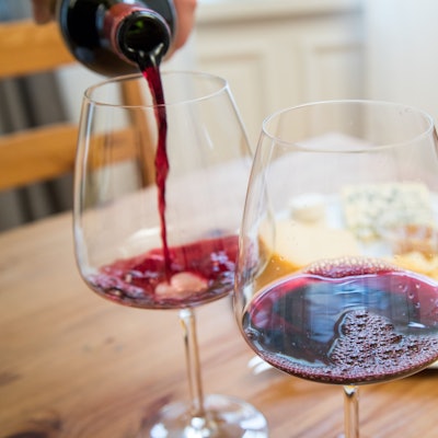 Im Vordergrund sehen zwei Rotweingläser, in eines wird gerade eingeschenkt. Im Hintergrund sieht man unscharf einen Teller mit Essen.