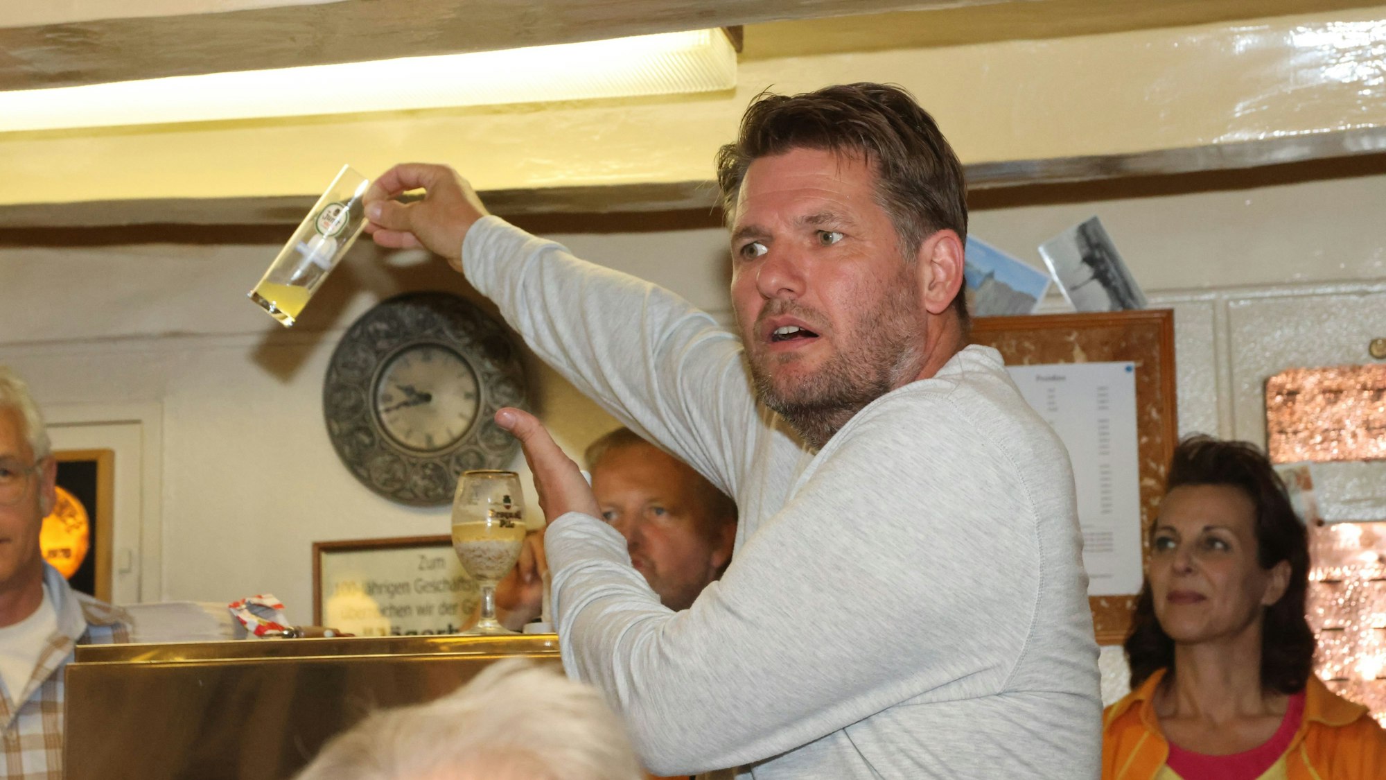 Ein Mann hält vor einem Publikum ein Bierglas in der Hand in die Höhe und zeigt darauf.