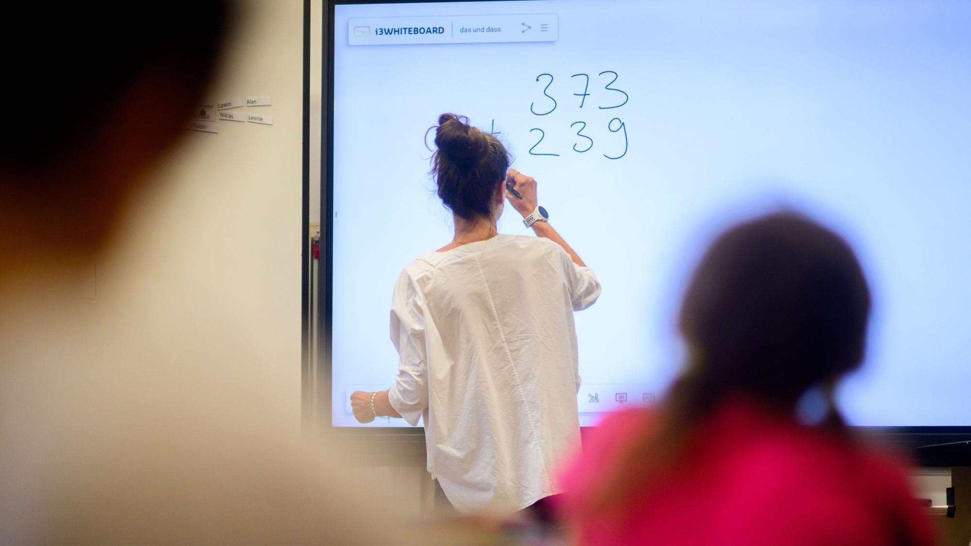 Ein Lehrerin schreibt eine Mathematikaufgabe auf eine digitale Schultafel im Klassenraum einer 4. Klasse einer Grundschule.