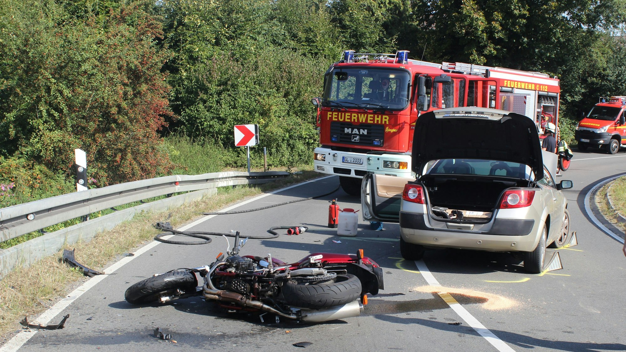 Ein schwer beschädigtes Motorrad liegt auf der Straße, dahinter stehen ein Auto sowie Einsatzfahrzeuge der Feuerwehr Kürten.