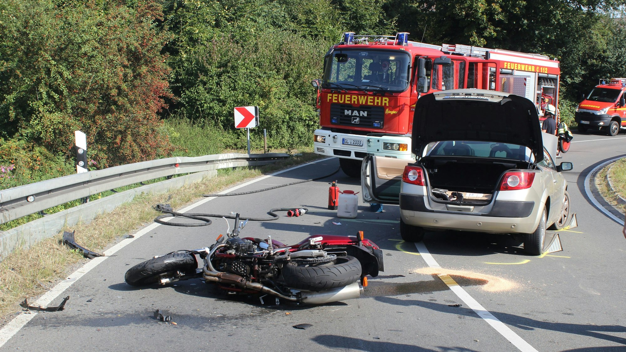 Ein Motorrad liegt auf der Fahrbahn, dahinter der Unfall-Pkw und zwei Einsatzfahrzeuge der Feuerwehr.