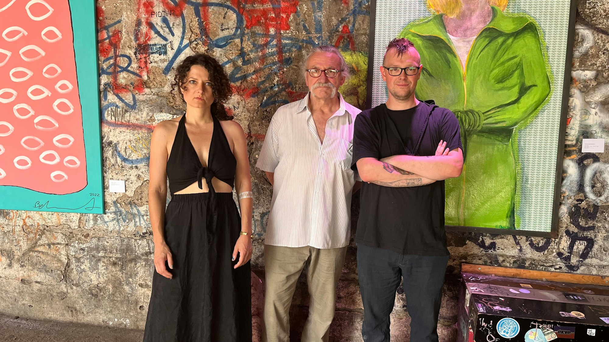 Eine Frau uns zwei Männer stehen vor einer bunt bemalten Betonwand, an der großformatige Bilder hängen.