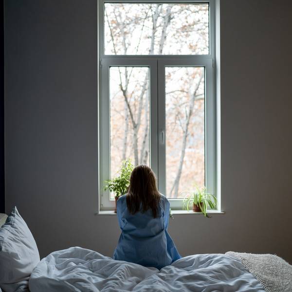 Eine Frau sitzt mit dem Rücken zur Kamera auf einem Bett und blickt aus dem Fenster.
