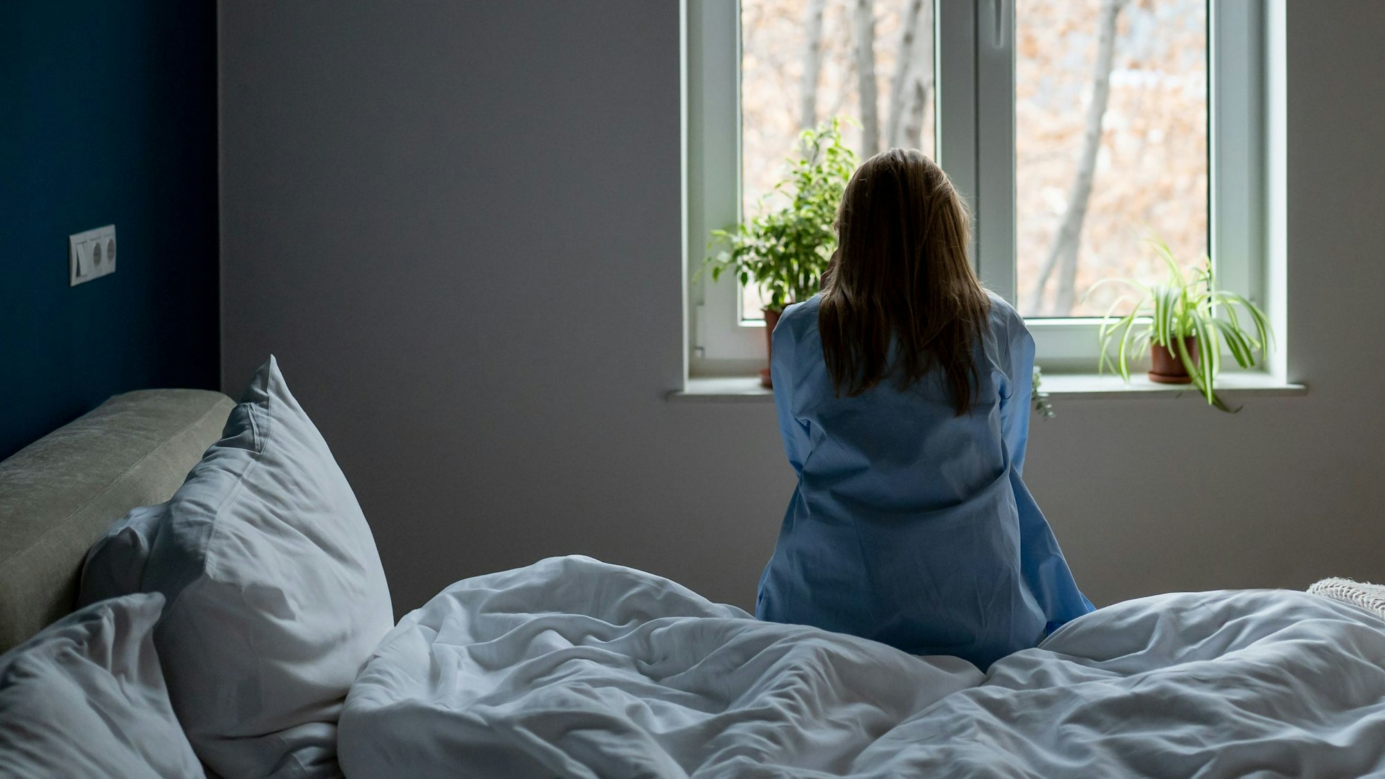 Eine Frau sitzt mit dem Rücken zur Kamera auf einem Bett und blickt aus dem Fenster.
