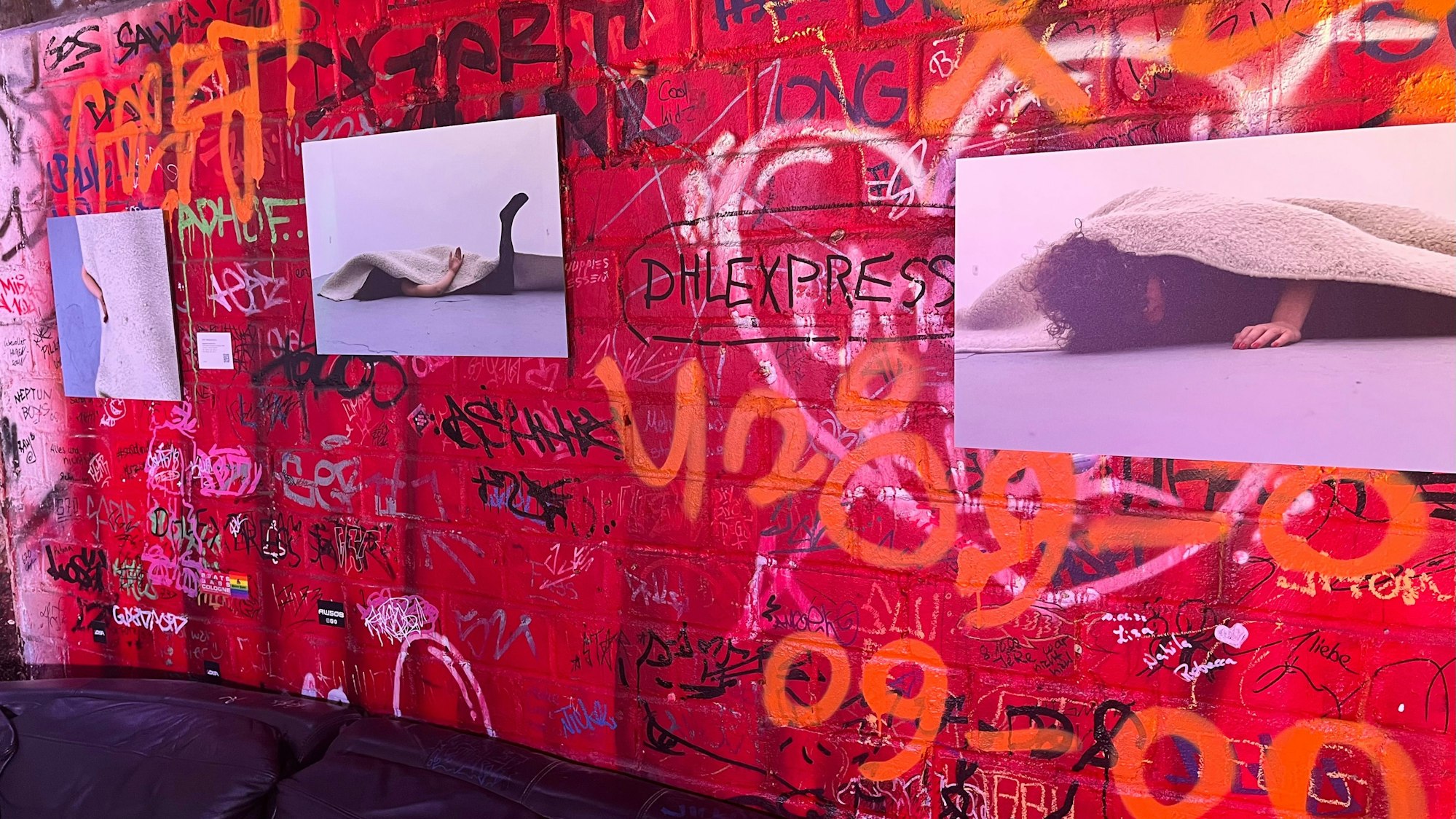 An einer roten bekritzelten Wand hängen Bilder von einer Frau, die halb unter einem Teppich liegt.