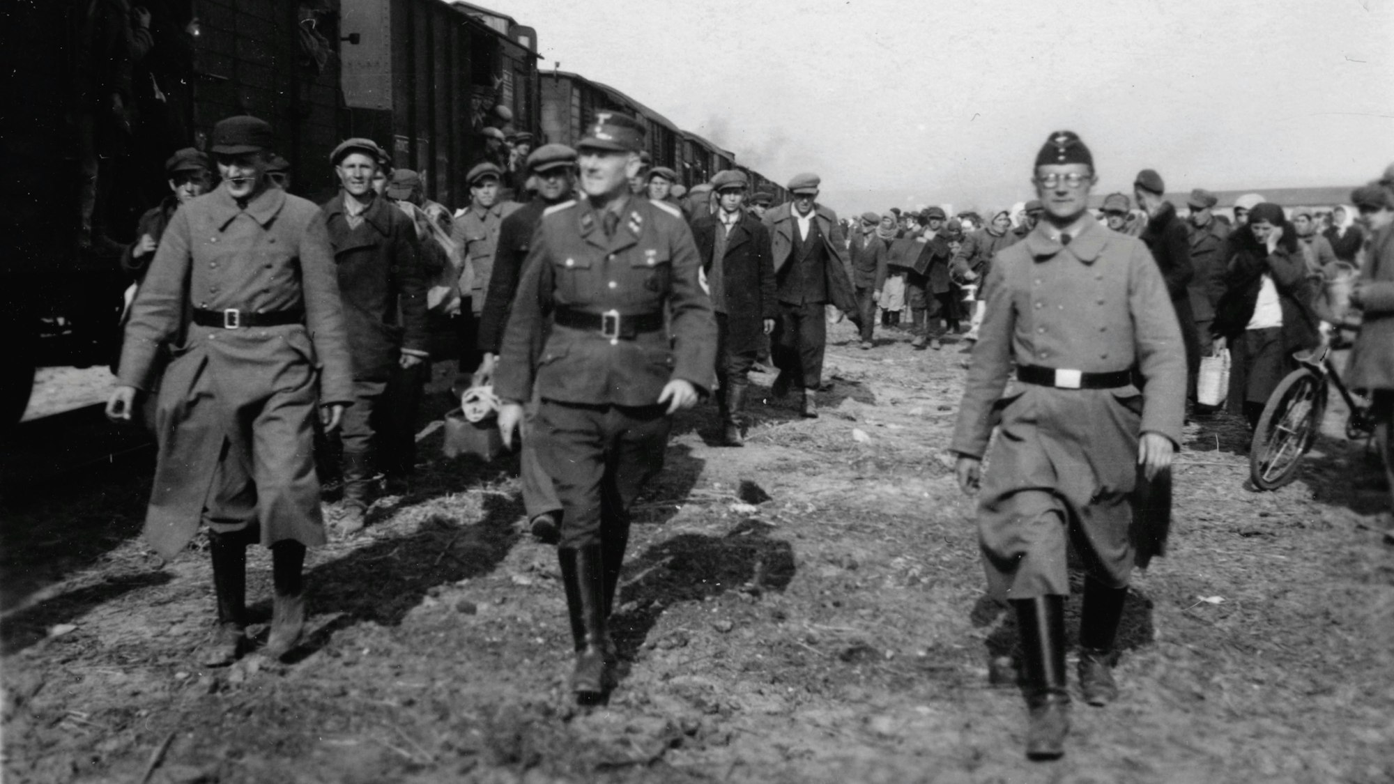 Das Bild zeigt drei deutsche Soldaten während des Zweiten Weltkriegs in Uniform. Im Hintergrund sind Zugwaggons und zahlreiche Zivilisten zu sehen.