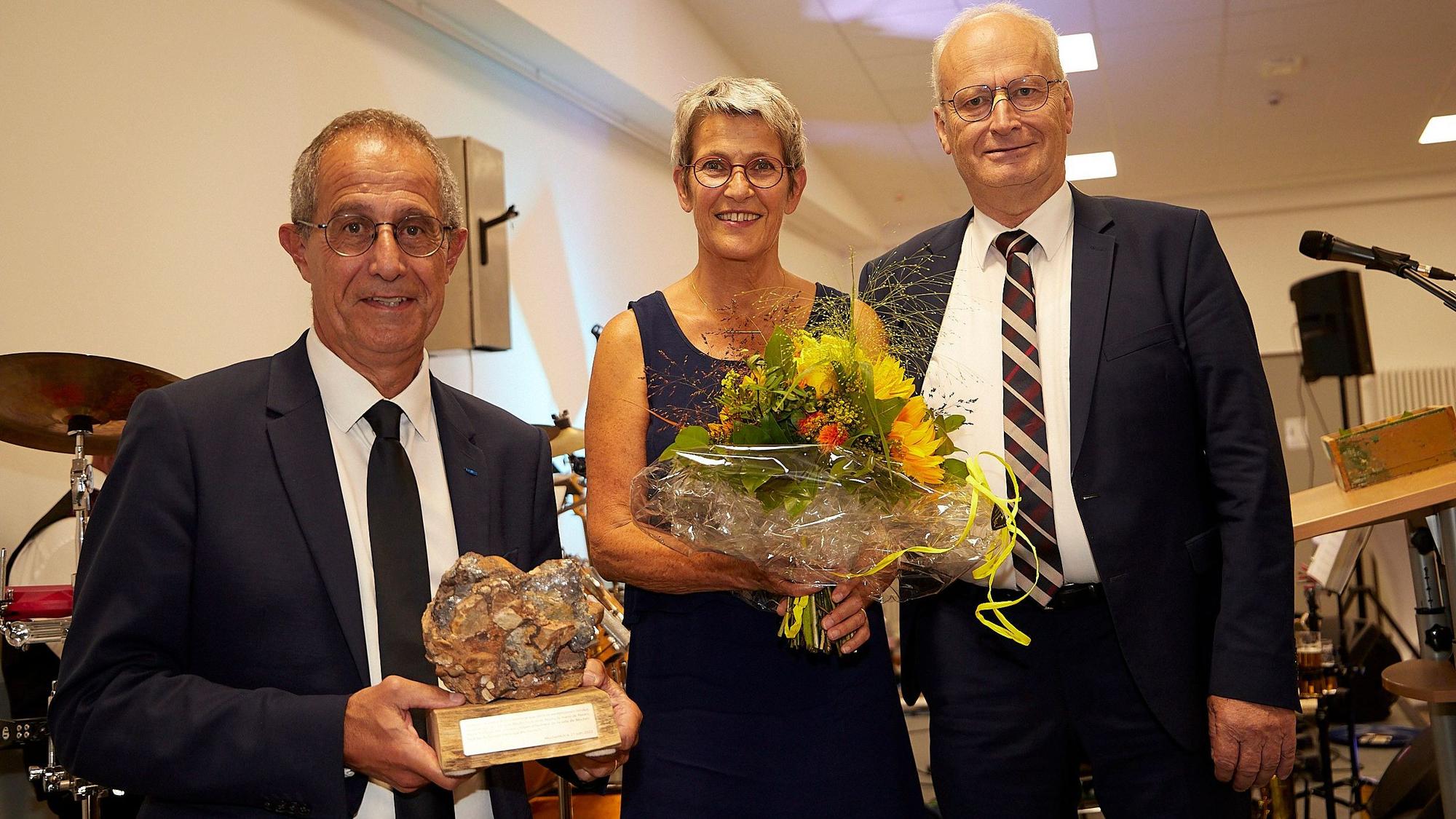 Zwei Männer und eine Frau, Dr. Hans-Peter Schick (r.) sowie Pierre und Juliane Combes, stehen nebeneinander. Die Männer tragen dunkle Anzüge. Der Mann links hält eine Bleiknolle in der Hand, die Frau einen Blumenstrauß.