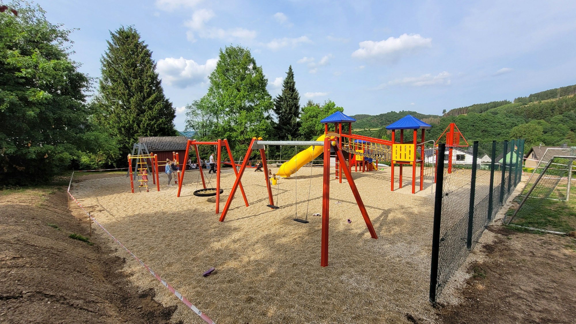 Rote Spielgeräte stehen auf einem Platz aus Kies. Zu sehen sind mehrere Schaukeln, eine gelbe Rutsche und Klettermöglichkeiten.