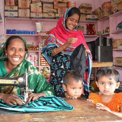 Eine junge Frau sitzt an einer Nähmaschine. Neben ihr stehen zwei Kinder.