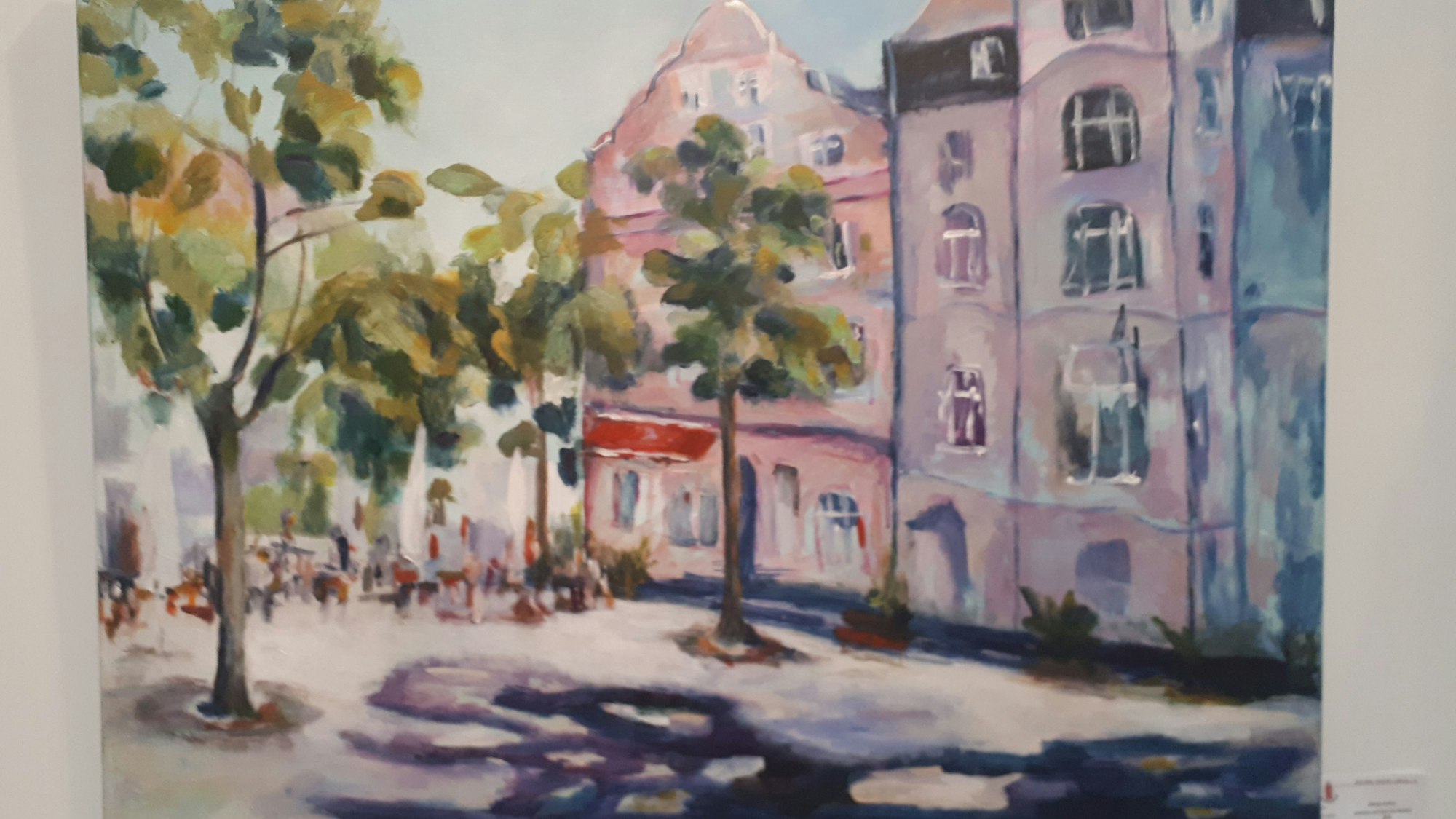 Ein Aquarell vom sommerlichen Schillplatz ist zu sehen. Menschen sitzen draußen an Café-Tischen. Bäume stehen vor den Häusern am beliebten Platz in Nippes.