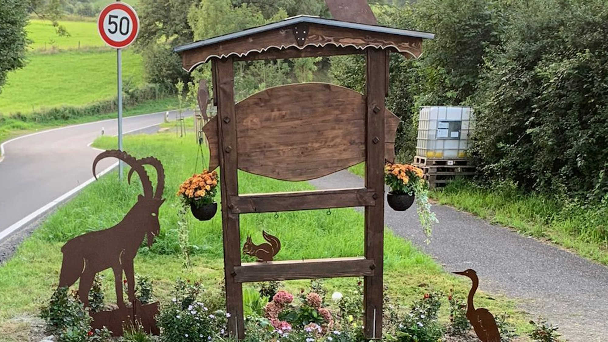 Das schön gestaltete Willkommensschild von Lanzenbach aus Holz n einem Blumenbeet mit fünf Tierfiguren aus Cortenstahl: Steinbock, Eule, Specht, Reiher und Eichhörnchen.