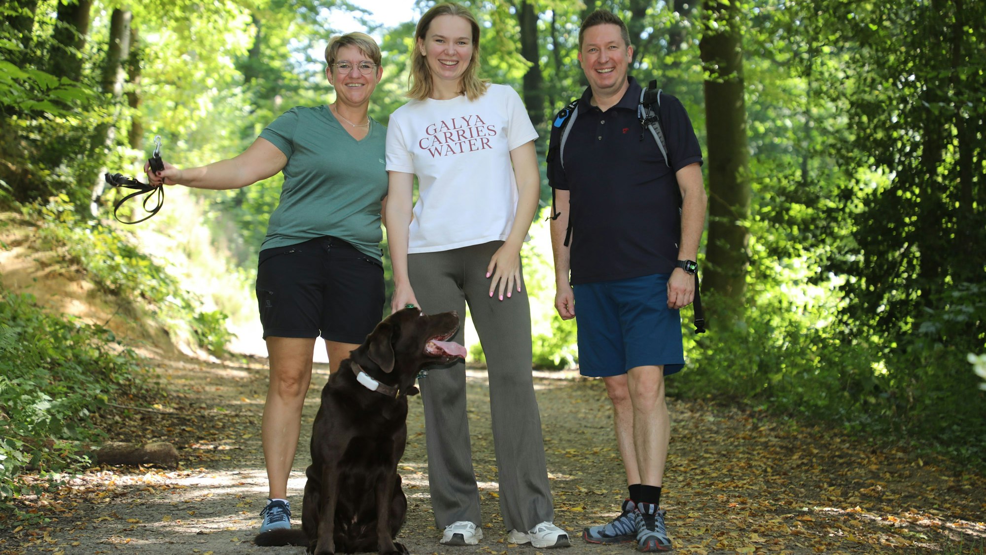 Drei Mnschen stehen mit einem Hund auf einem Waldweg.