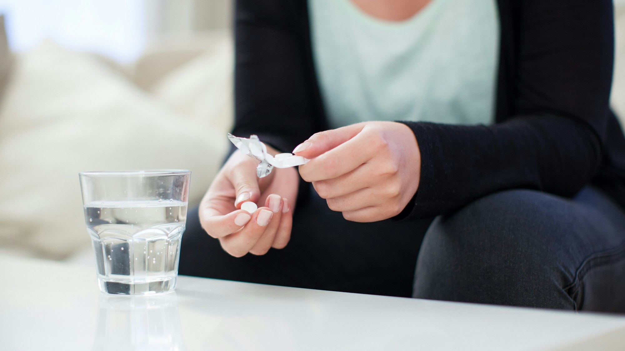 Eine Person nimmt eine Schmerztablette aus der Verpackung. Vor ihr steht ein Glas Wasser auf dem Tisch.