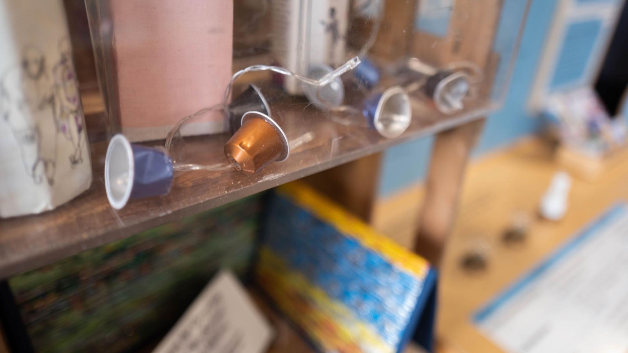Das Bild zeigt den Inhalt einer Vitrine. Zu sehen sind benutzte Kaffeekapseln, die mit einer Lichterkette verbunden sind.